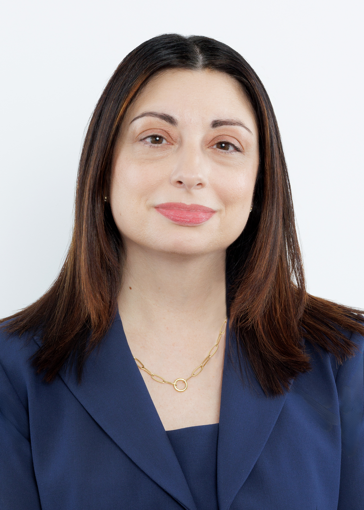 A woman with straight brown hair wearing a navy blazer, gold chain necklace, and neutral makeup, smiling slightly against a plain white background.