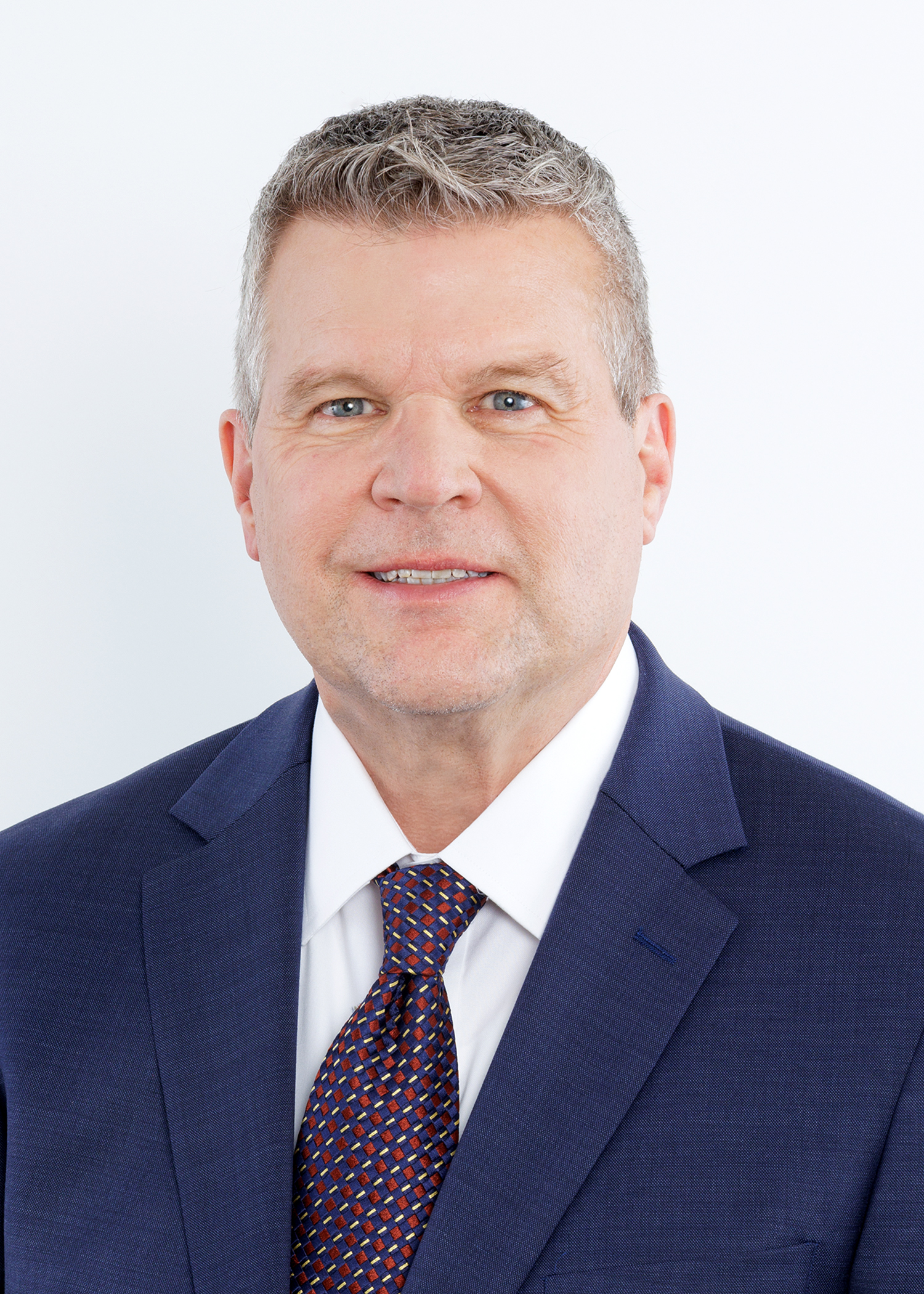 A middle-aged man with short gray hair, wearing a navy blue suit, white shirt, and patterned tie, smiles slightly against a plain white background.