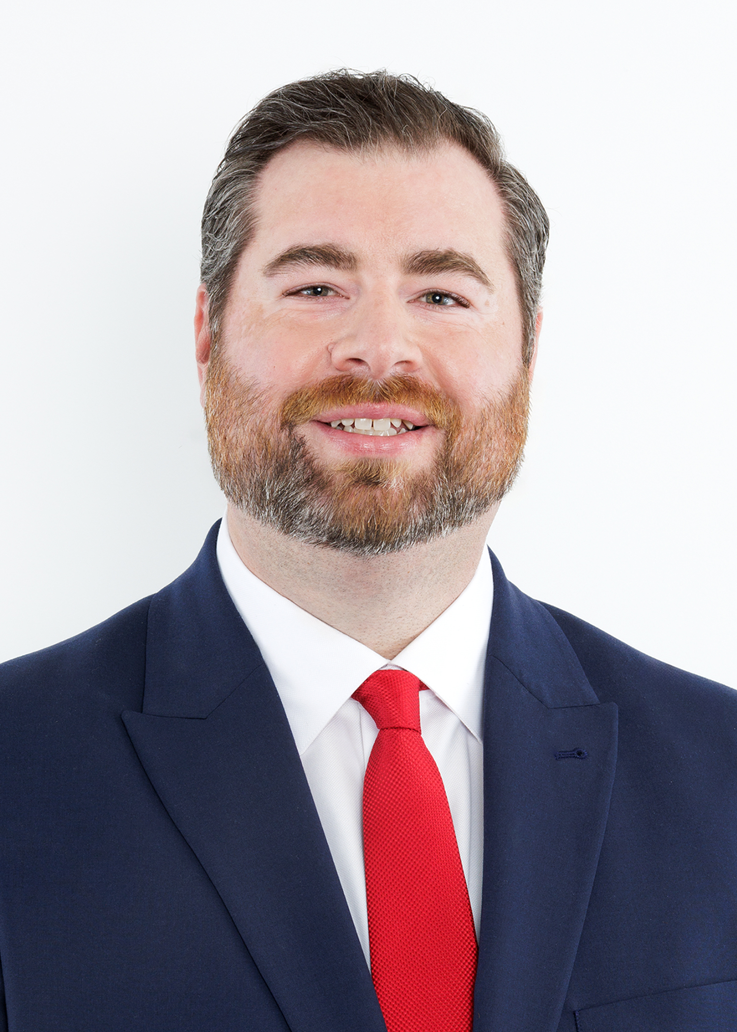 A man with short brown hair and a beard, wearing a navy blue suit, white shirt, and red tie, smiles in front of a plain white background.