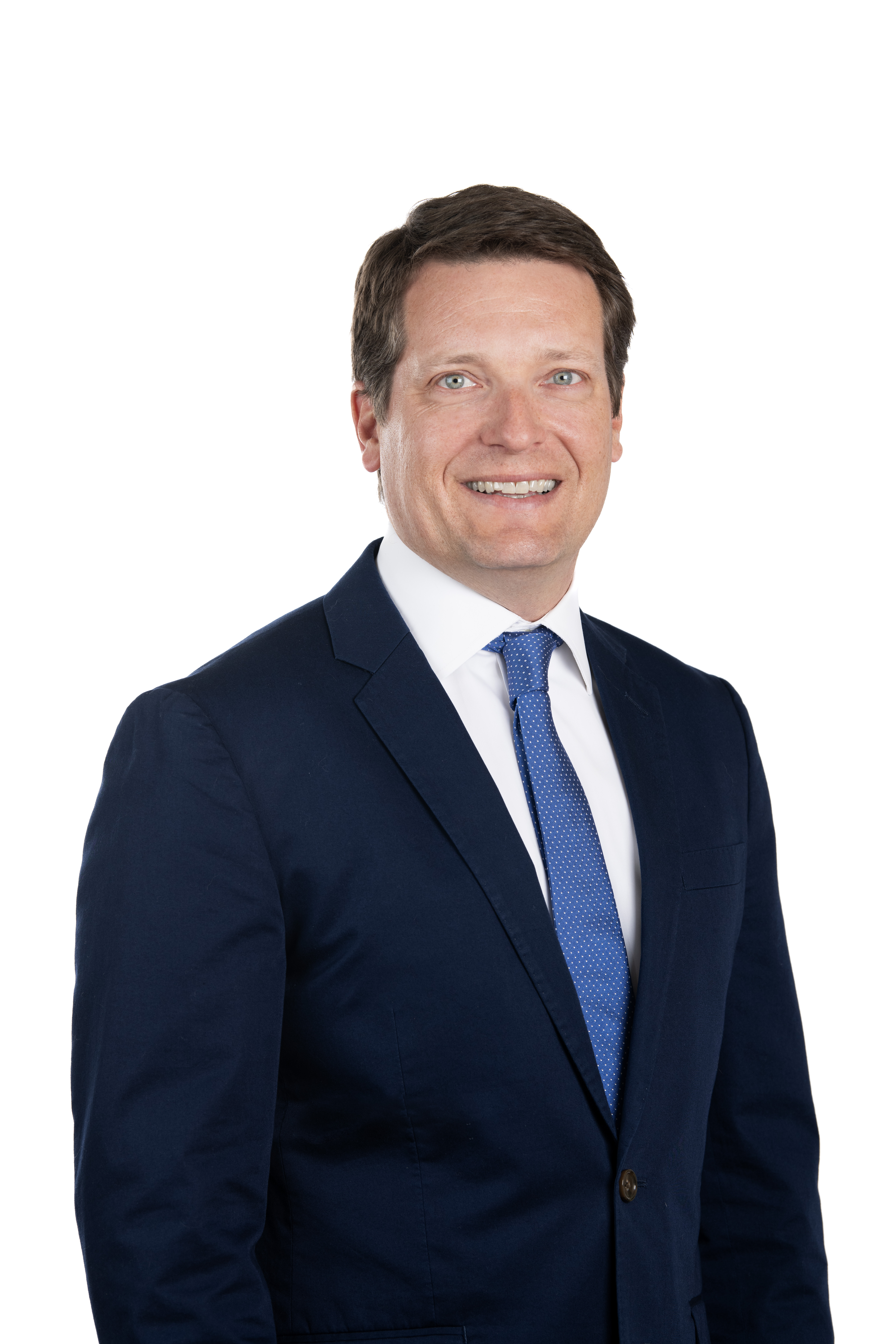 A man in a navy suit, white shirt, and blue tie smiles while standing against a plain white background.