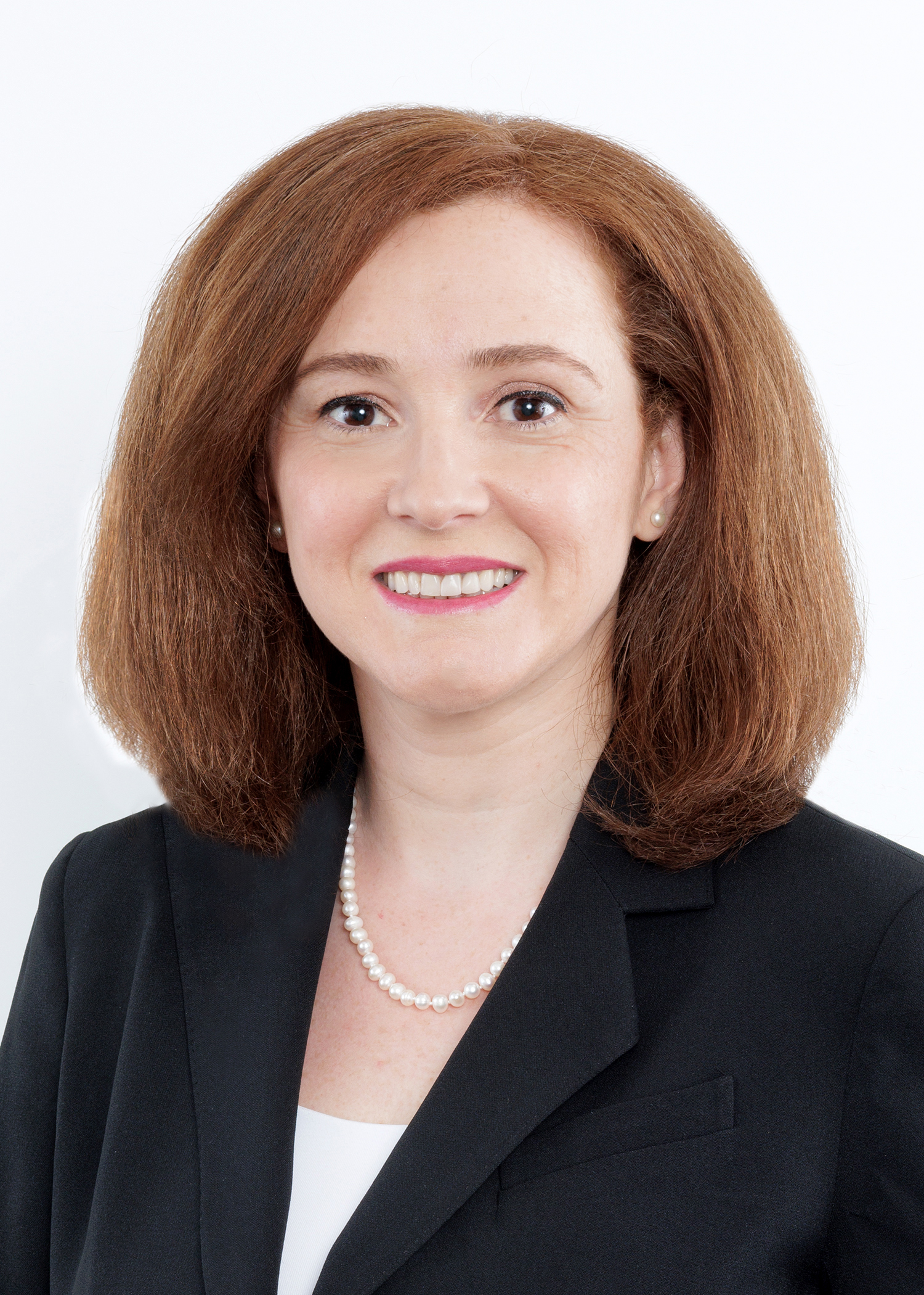 A woman with shoulder-length reddish-brown hair, wearing a black blazer, white top, pearl necklace, and stud earrings, smiles at the camera against a plain white background.