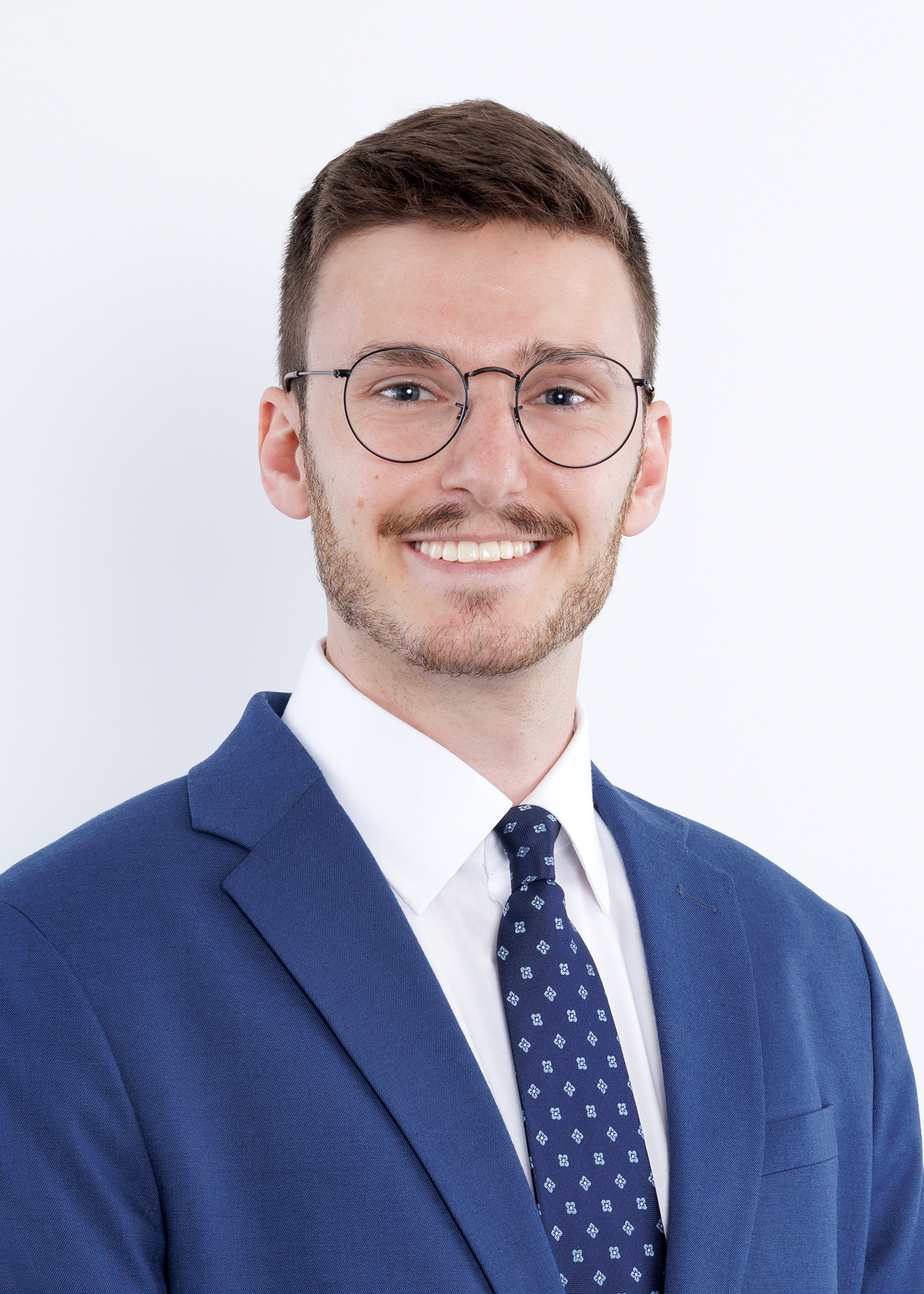 JustinCarter A young man with short brown hair, glasses, and light stubble smiles while wearing a blue suit, white shirt, and patterned navy tie against a plain white background.