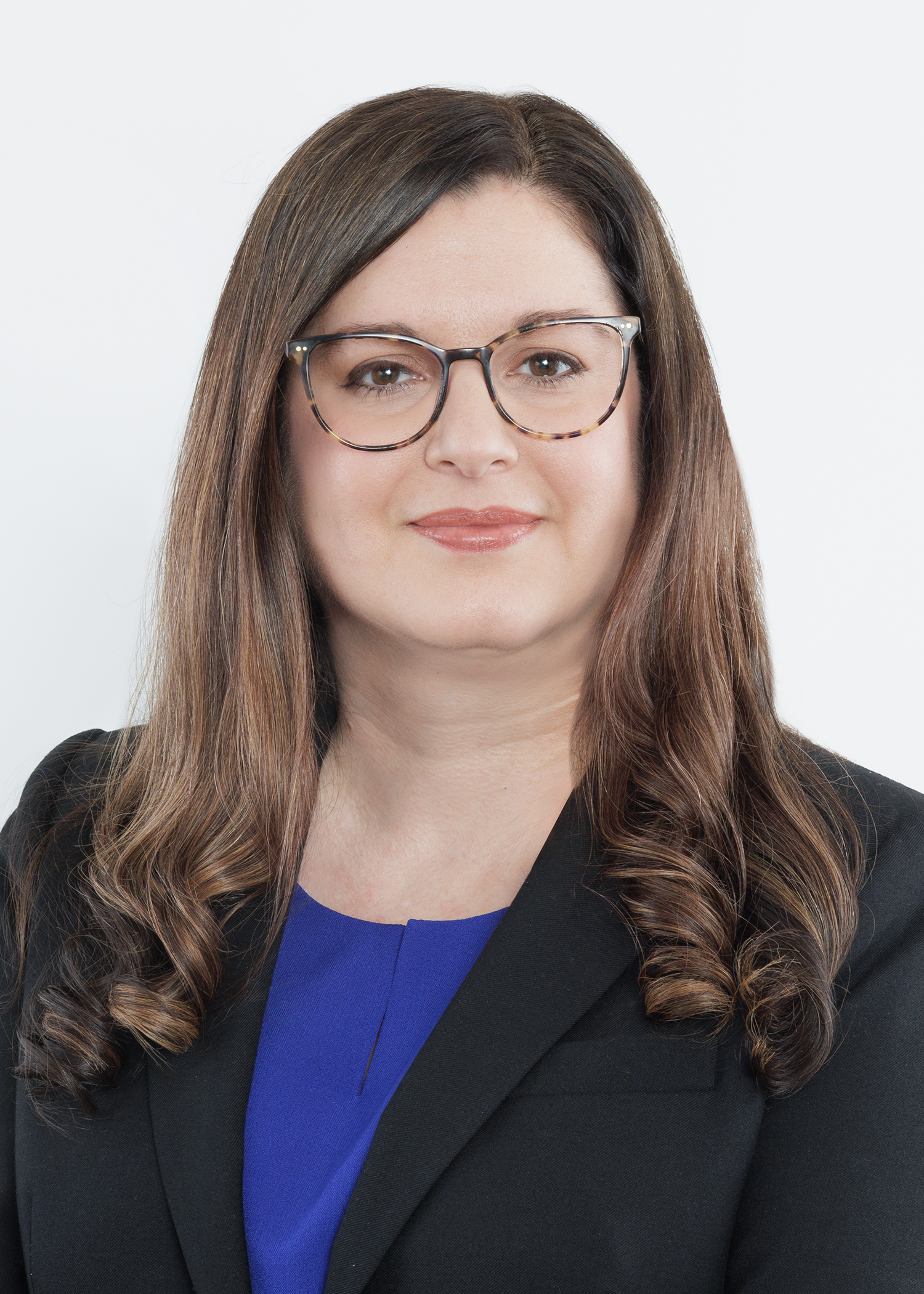 Woman with long brown hair and glasses, wearing a black blazer over a blue top, smiling slightly against a plain white background.
