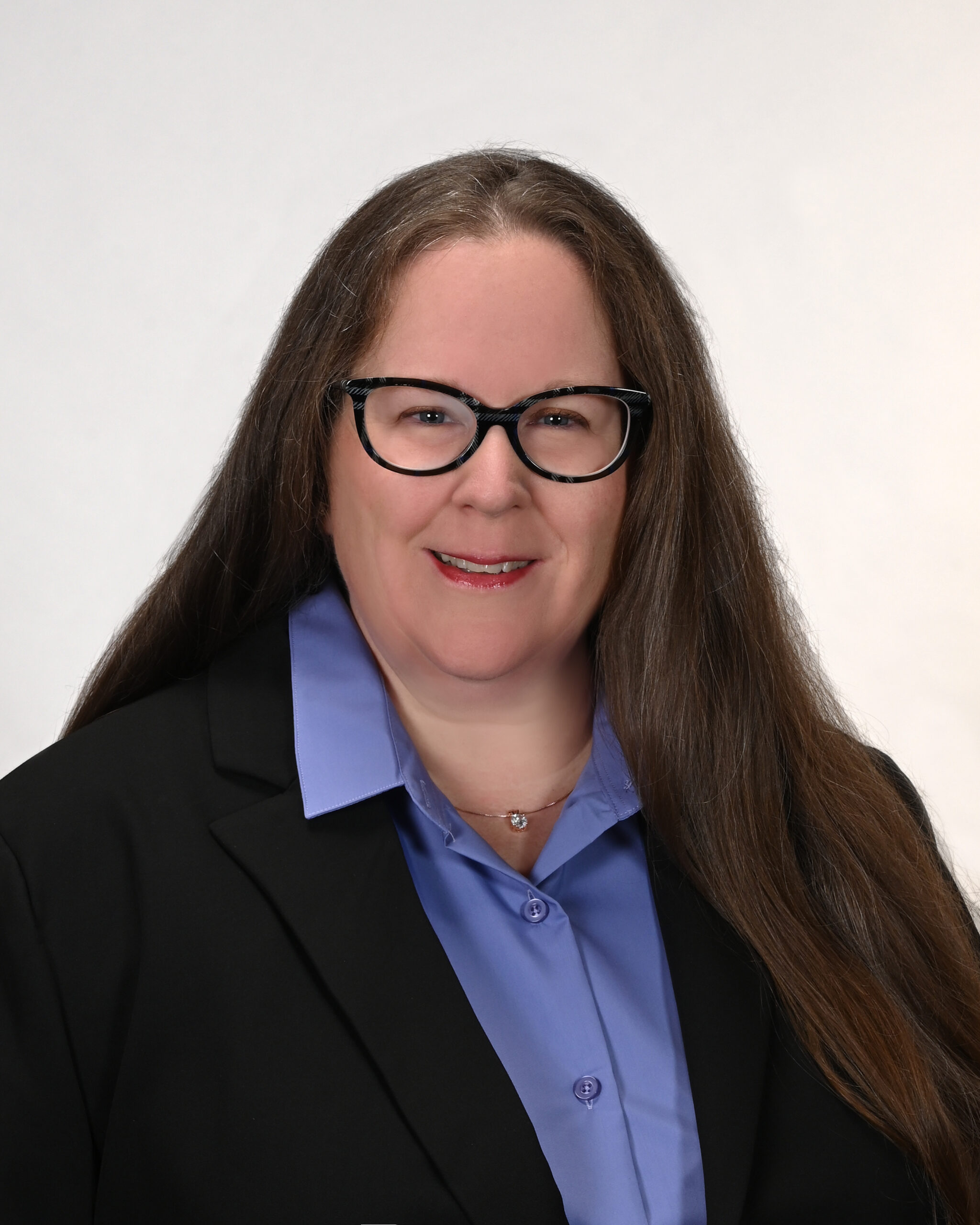 A woman with long brown hair and glasses smiles while wearing a black blazer over a light blue collared shirt, standing against a plain light background.