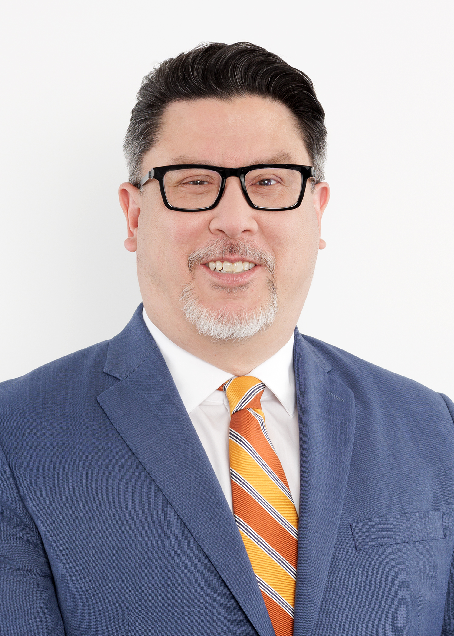 A man wearing glasses, a blue suit, white shirt, and an orange striped tie smiles in front of a plain white background.